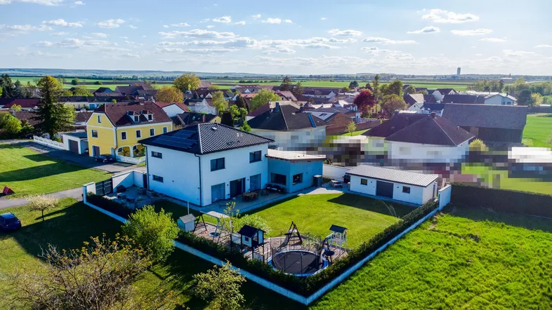 Luftaufnahme eines modernen Hauses mit großem Garten, Spielplatz und Photovoltaikanlage, umgeben von Feldern.