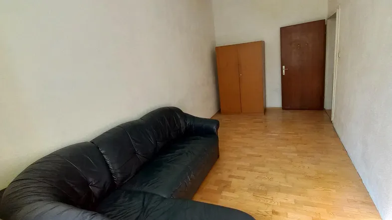 Geräumiges Wohnzimmer mit dunklem Ledersofa, hellem Parkettboden und einfacher Einrichtung.