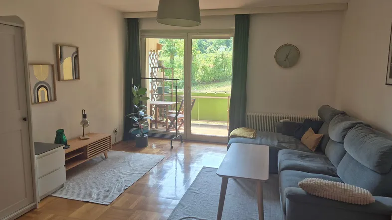 Helles Wohnzimmer mit Parkettboden, gemütlichem Sofa und Zugang zu einem Balkon mit Grünblick.