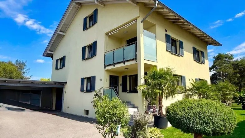 Gelbes Einfamilienhaus mit blauen Fensterläden, Balkon und gepflegtem Vorgarten unter blauem Himmel.