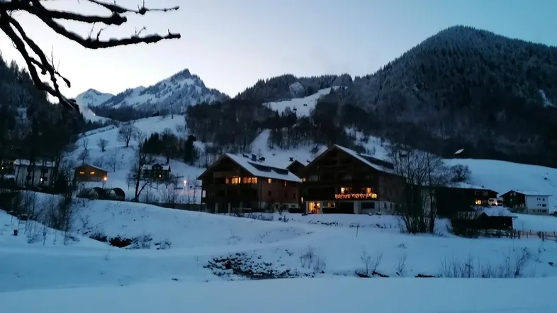 Winterliche Abendstimmung mit beleuchteten Chalet-Gebäuden und schneebedeckten Bergen im Hintergrund.