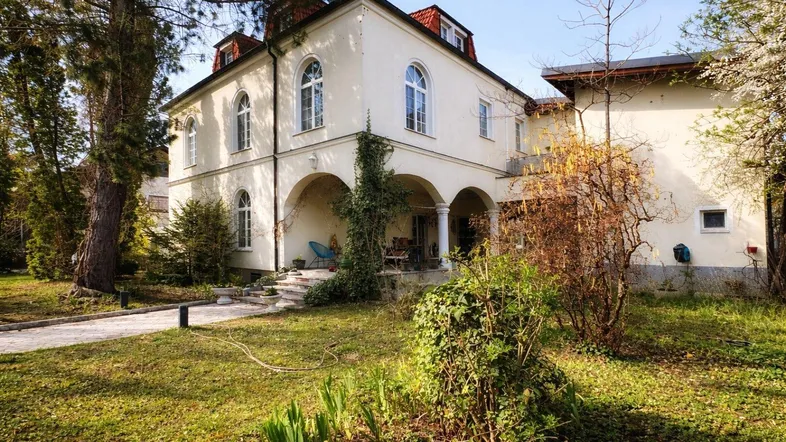 Seitenansicht der Villa mit großem Garten, altem Baumbestand und überdachtem Eingangsbereich.