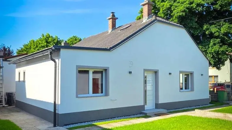 Modernes Einfamilienhaus mit weißer Fassade, grauem Sockel und gepflegtem Rasen unter blauem Himmel.