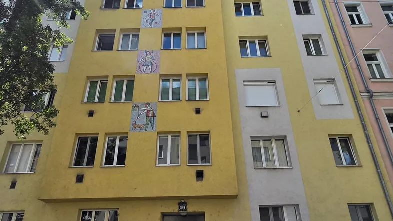 Gelbe Fassade eines Mehrfamilienhauses mit mehreren Fenstern und dekorativen Elementen unter blauem Himmel.