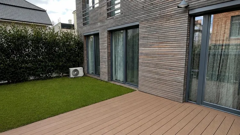 Moderne Holzterrasse mit angrenzendem Rasenbereich und Fassade des Gebäudes in Holzoptik.