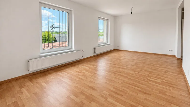 Helles, leeres Zimmer mit Laminatboden, weißen Wänden und Blick ins Grüne durch die Fenster.