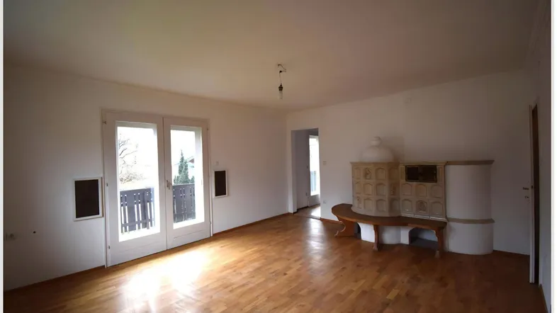 Helles Wohnzimmer mit Holzboden, traditionellem Kachelofen und Zugang zu einem Balkon.