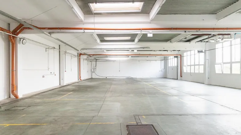 Weitläufige Industriehalle mit Betonboden, sichtbaren Leitungen und großen Fenstern für viel Tageslicht.