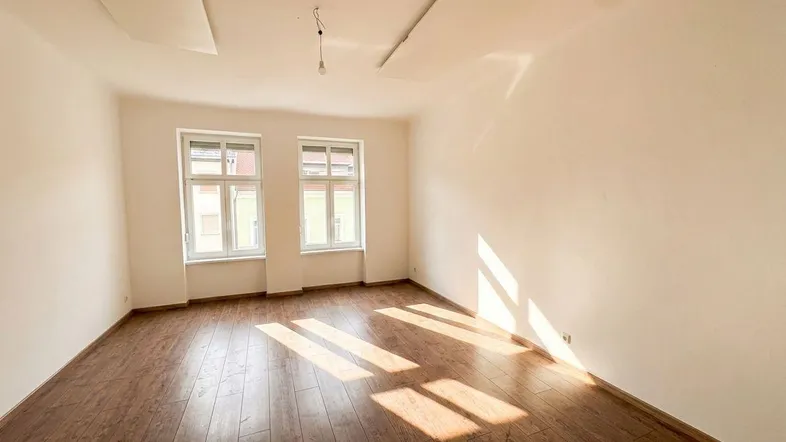 Helles Zimmer mit Holzboden und zwei Fenstern, durch die Sonnenlicht einfällt.