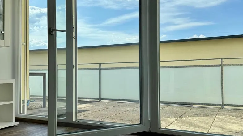 Heller Innenraum mit großen Fenstern und direktem Zugang zu einer geräumigen Terrasse unter blauem Himmel.