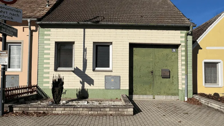 Älteres Gebäude mit grüner Garagentür und gepflastertem Vorplatz unter blauem Himmel.