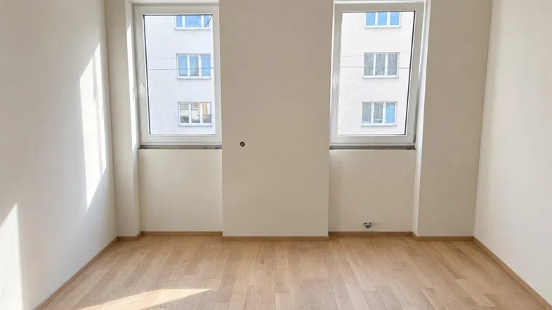 Heller Raum im Rohbau mit Parkettboden und zwei Fenstern, die auf ein gegenüberliegendes Gebäude blicken.