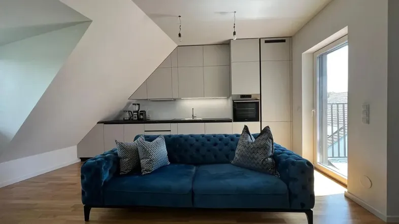 Helles Wohnzimmer mit blauer Couch, offener Küche und Holzboden unter einer Dachschräge.