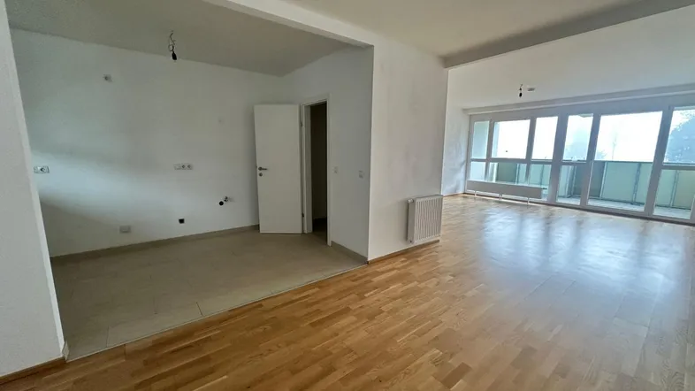 Geräumiger offener Wohnbereich mit Parkettboden, großen Fenstern und Zugang zu einem Balkon.