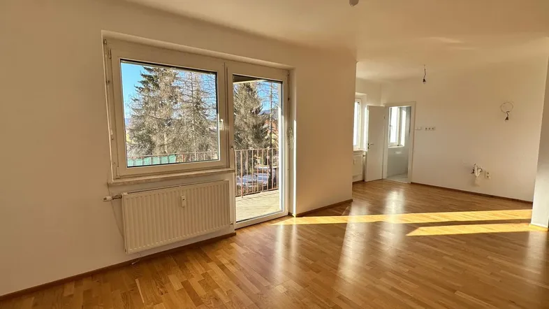 Helles Wohnzimmer mit Parkettboden und Zugang zu einem kleinen Balkon, durchflutet von Sonnenlicht.