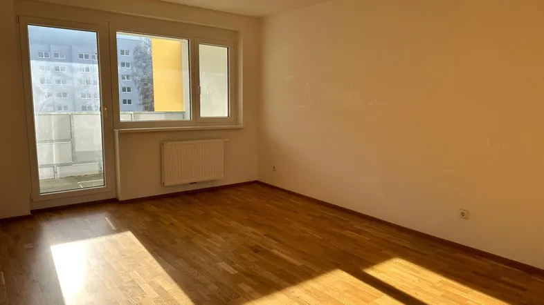 Helles, leeres Wohnzimmer mit Parkettboden und Zugang zu einem Balkon.