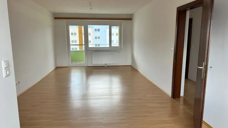 Helles, leeres Wohnzimmer mit Laminatboden und Zugang zu einem Balkon, mit Blick auf andere Gebäude.