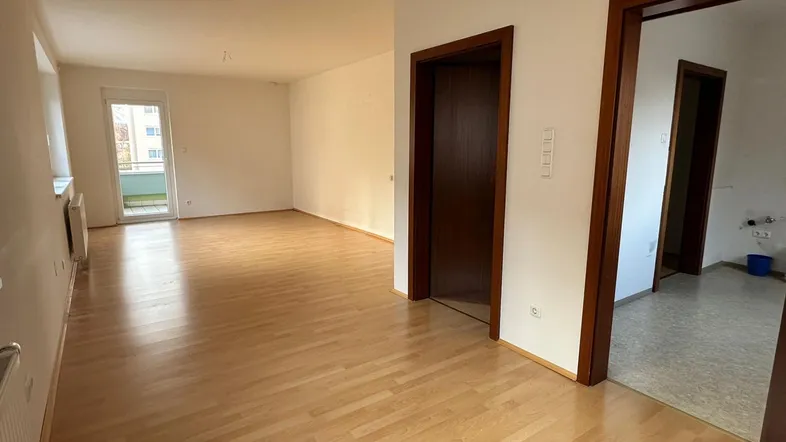 Helles, leeres Wohnzimmer mit Laminatboden und Zugang zu einem kleinen Balkon.
