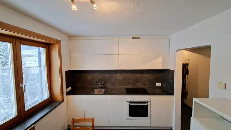 Kleine, moderne Küche mit weißen Schränken, dunkler Arbeitsplatte und Fensterblick ins Freie.
