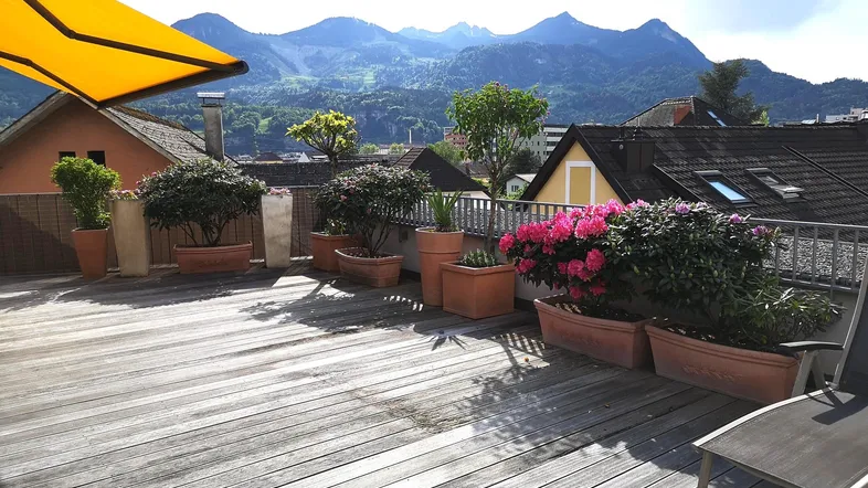 Geräumige Dachterrasse mit Holzboden, vielen Pflanzen, Sonnenmarkise und beeindruckendem Bergblick.