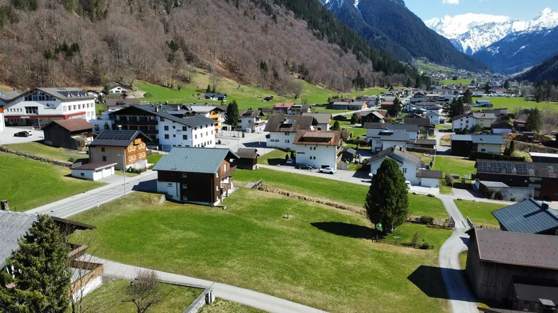 Luftaufnahme eines Dorfes in den Bergen mit grünen Wiesen und traditionellen Häusern unter blauem Himmel.