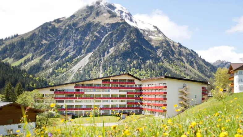 Großes Gebäude mit vielen Balkonen vor einer beeindruckenden Bergkulisse im Sommer.