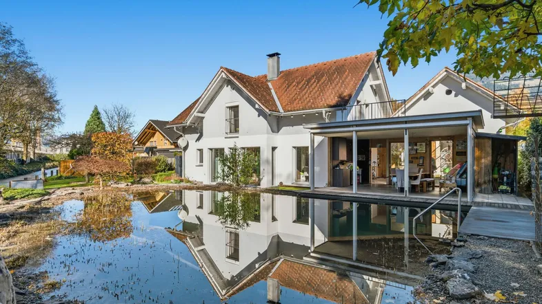Weißes Haus mit Terrakotta-Dach und natürlichem Schwimmteich, umgeben von Herbstbäumen.