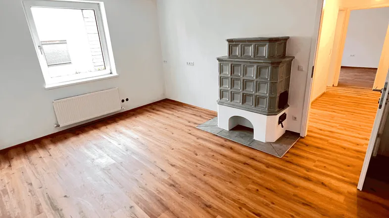 Helles Zimmer mit Holzboden und traditionellem Kachelofen, bereit zur individuellen Gestaltung.