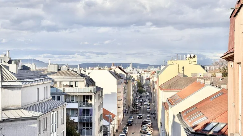 Blick über eine belebte Stadtstraße mit Gebäuden und Bergen im Hintergrund unter bewölktem Himmel.