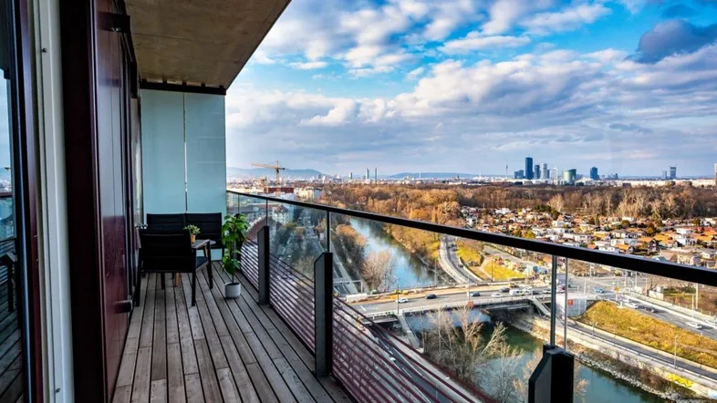 Geräumiger Balkon mit Holzboden, Glasgeländer und weitem Blick über die Stadt und den Fluss.