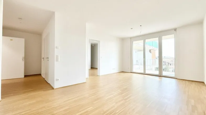 Helles, leeres Wohnzimmer mit Parkettboden und großen Fenstern, die Zugang zu einem Balkon bieten.