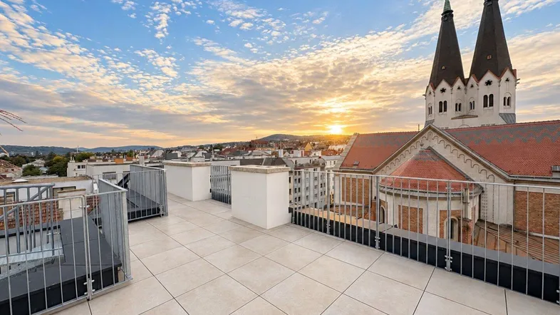 Geräumige Dachterrasse mit Fliesenboden und weitem Blick über die Stadt und eine Kirche bei Sonnenuntergang.