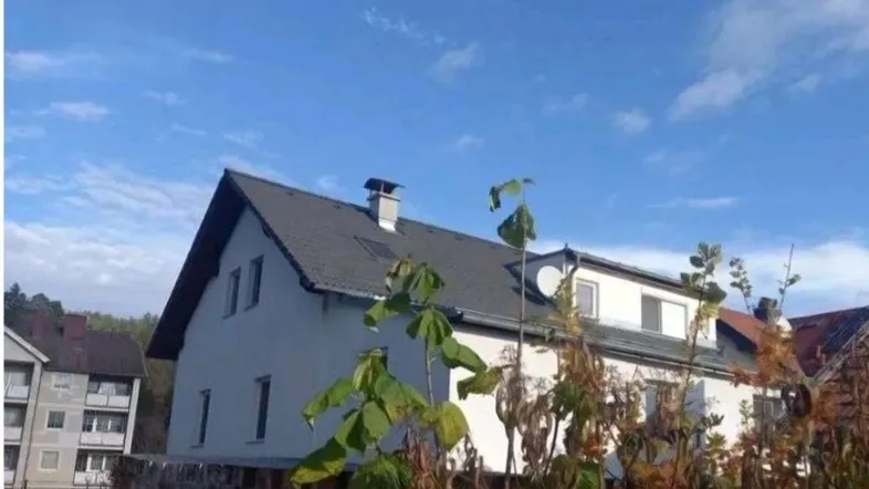 Hausfassade mit Satteldach und blauer Himmel im Hintergrund, umgeben von Bäumen.