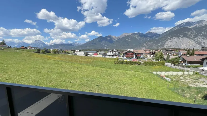 Panoramablick von einem Balkon über grüne Felder auf ein Dorf und die umliegende Berglandschaft.
