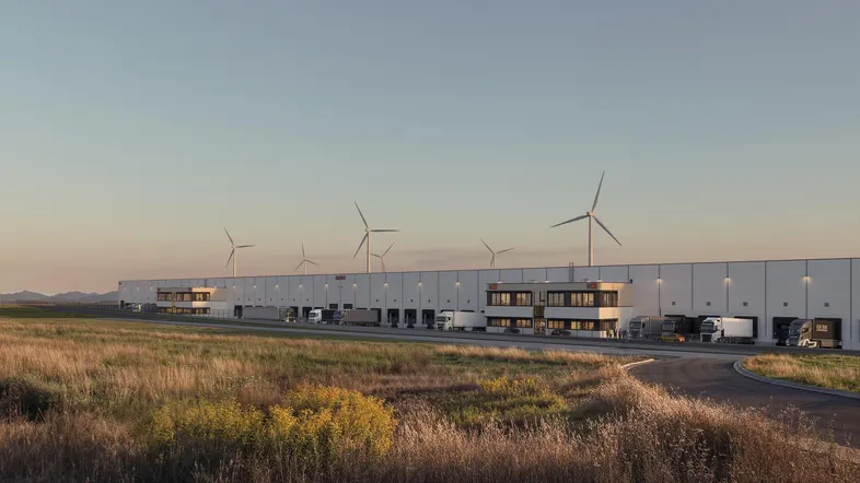 Großes Logistikzentrum mit mehreren Laderampen und Windkraftanlagen im ländlichen Umfeld.