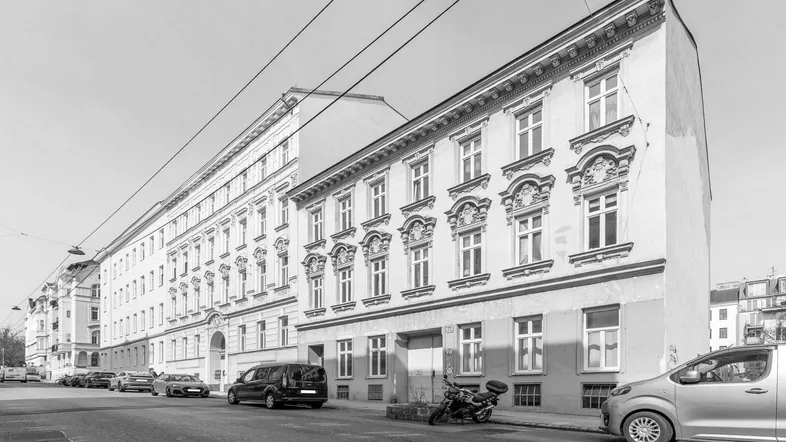 Schwarz-Weiß-Aufnahme einer historischen Gebäudefassade entlang einer städtischen Straße mit parkenden Autos.
