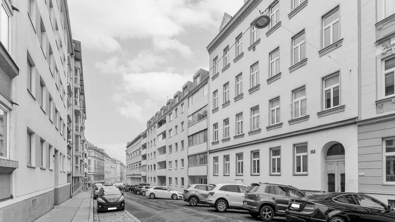 Schwarz-Weiß-Aufnahme einer städtischen Straße mit parkenden Autos und mehrstöckigen Gebäuden.