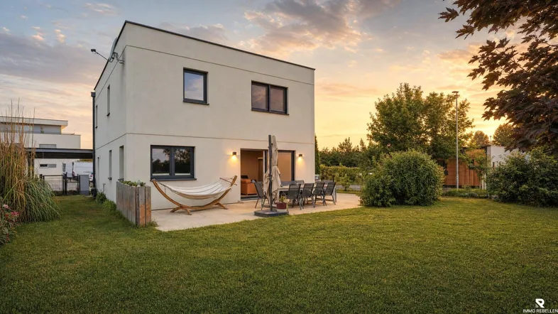Modernes Einfamilienhaus mit großer Rasenfläche und möblierter Terrasse im Sonnenuntergang.