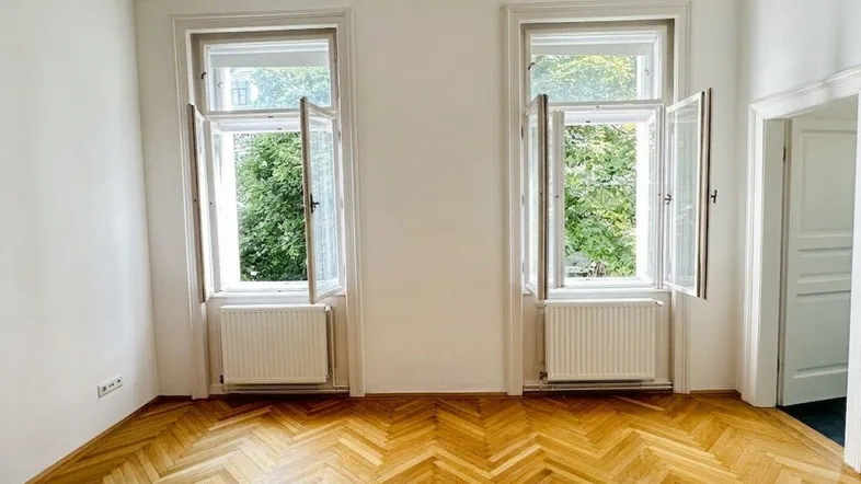 Helles Zimmer mit Fischgrätparkett und zwei großen Fenstern, die einen Blick ins Grüne bieten.