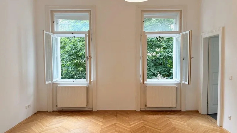 Helles Zimmer mit Fischgrätparkett und Blick ins Grüne durch zwei große Fenster.