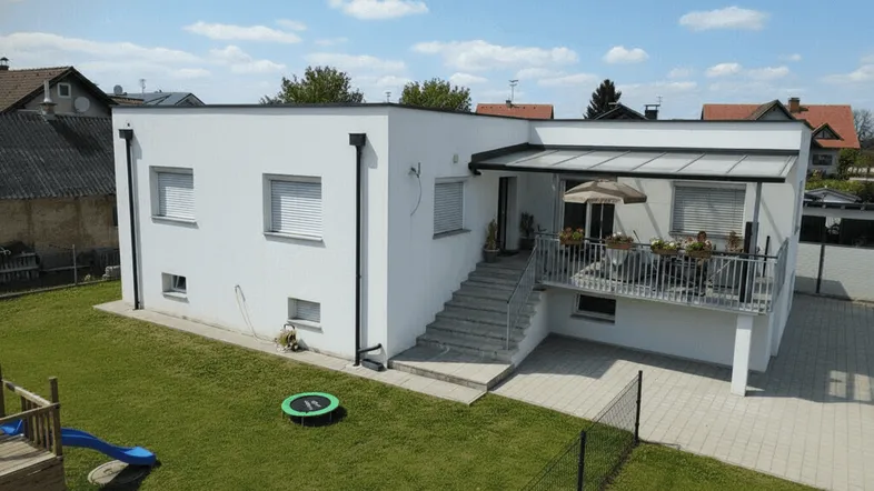 Modernes weißes Haus mit Flachdach, großem Garten und überdachter Terrasse mit Sitzbereich.