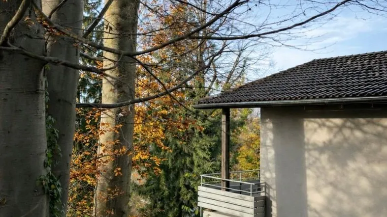 Teilansicht des Hauses mit kleinem Balkon, umgeben von Bäumen und herbstlichem Laub.