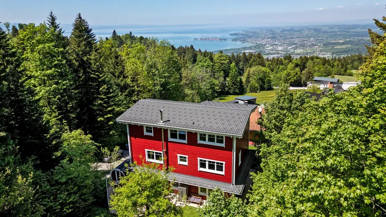 Panoramablick auf das rote Haus, umgeben von Wald, mit See- und Stadtblick in der Ferne.