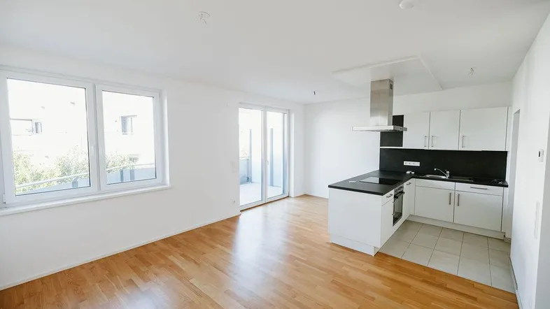 Heller offener Wohnbereich mit integrierter Küche, Parkettboden und Zugang zum Balkon.