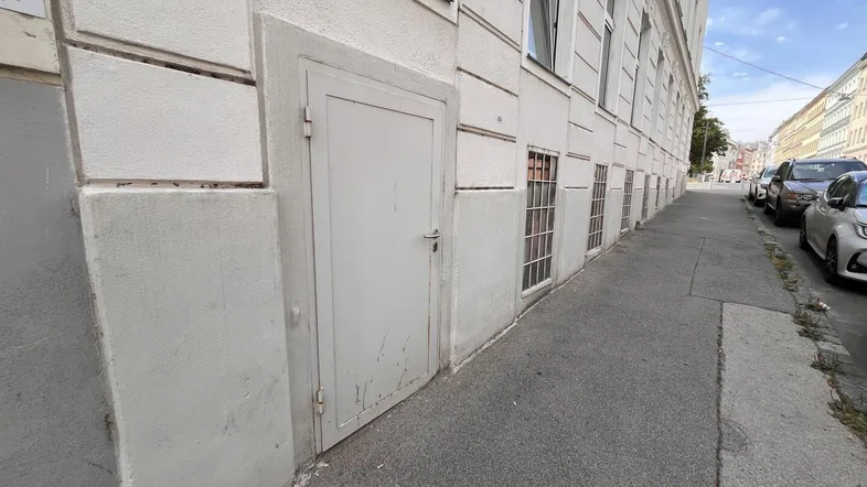 Seitenansicht eines Gebäudes mit einer einfachen Tür und vergitterten Kellerfenstern entlang des Bürgersteigs.