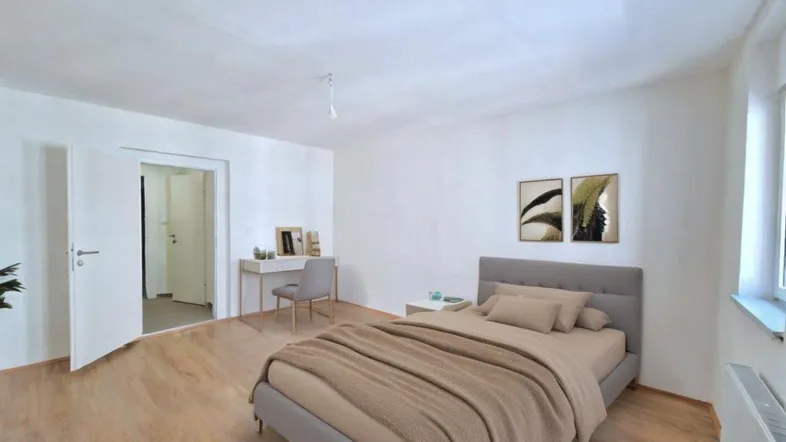 Virtuell inszeniertes Schlafzimmer mit Holzboden, weiß gestrichenen Wänden und einem kleinen Arbeitsbereich.