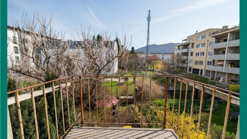 Sonniger Balkon mit Blick auf einen grünen Garten, umliegende Gebäude und entfernte Berge.