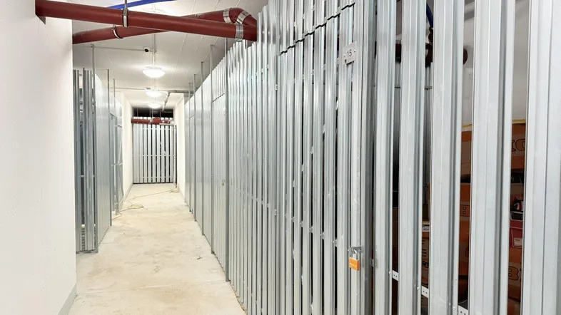Langer Flur im Kellerbereich mit zahlreichen individuellen Lagerabteilen aus Metall und sichtbaren Rohren an der Decke.