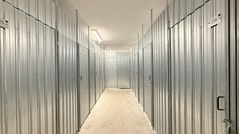 Langer Gang mit zahlreichen individuellen Lagerabteilen aus Metall und Betonboden, beleuchtet von Deckenleuchten.