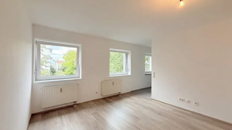 Helles, leeres Zimmer mit Laminatboden und zwei Fenstern, die einen grünen Ausblick bieten.
